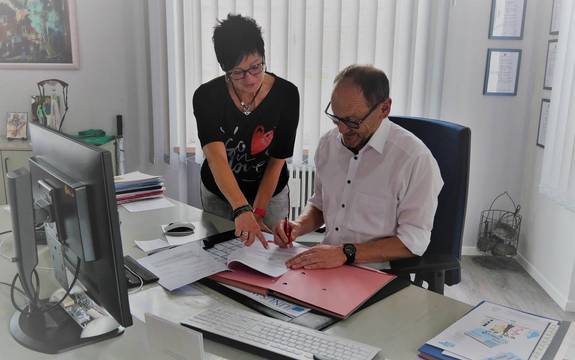Bürgermeister Markus Fuchs und Susanne Dour (Leiterin Gemeindekasse), stehen am Schreibtisch und unterzeichnen Vertrag